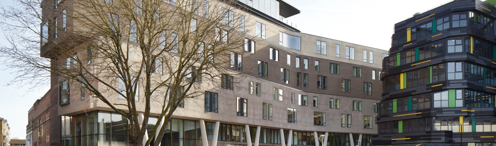 Grey curving building with students walking in front and small patch of grass. Clear blue sky. 