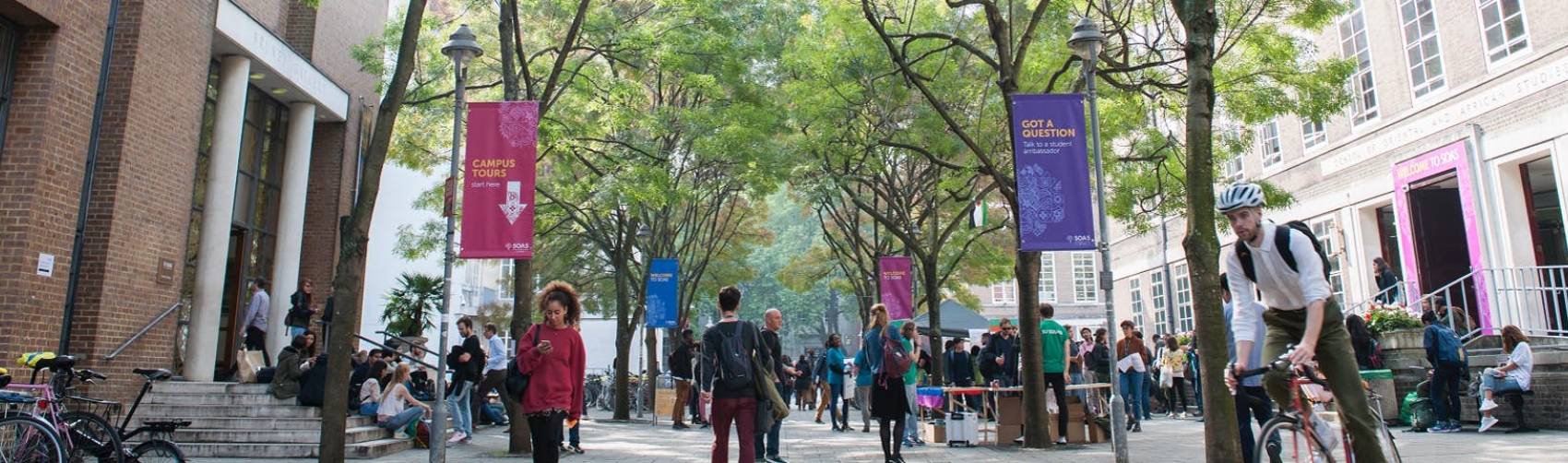 Landscape of building at left, people walking in foreground of SOAS buildings. Within concourse. 