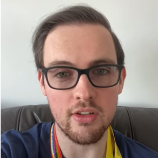 Gentlemen with black glasses wearing a rainbow lanyard and blue t-shirt. Sat on a sofa. He is mid sentence.
